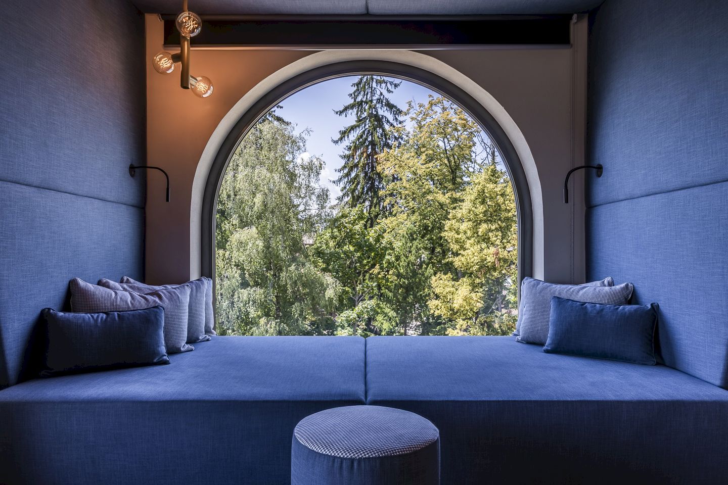 Posto finestra nella Lorenzi Suite con divano blu, cuscini e grande finestra arrotondata che si affaccia sugli alberi.