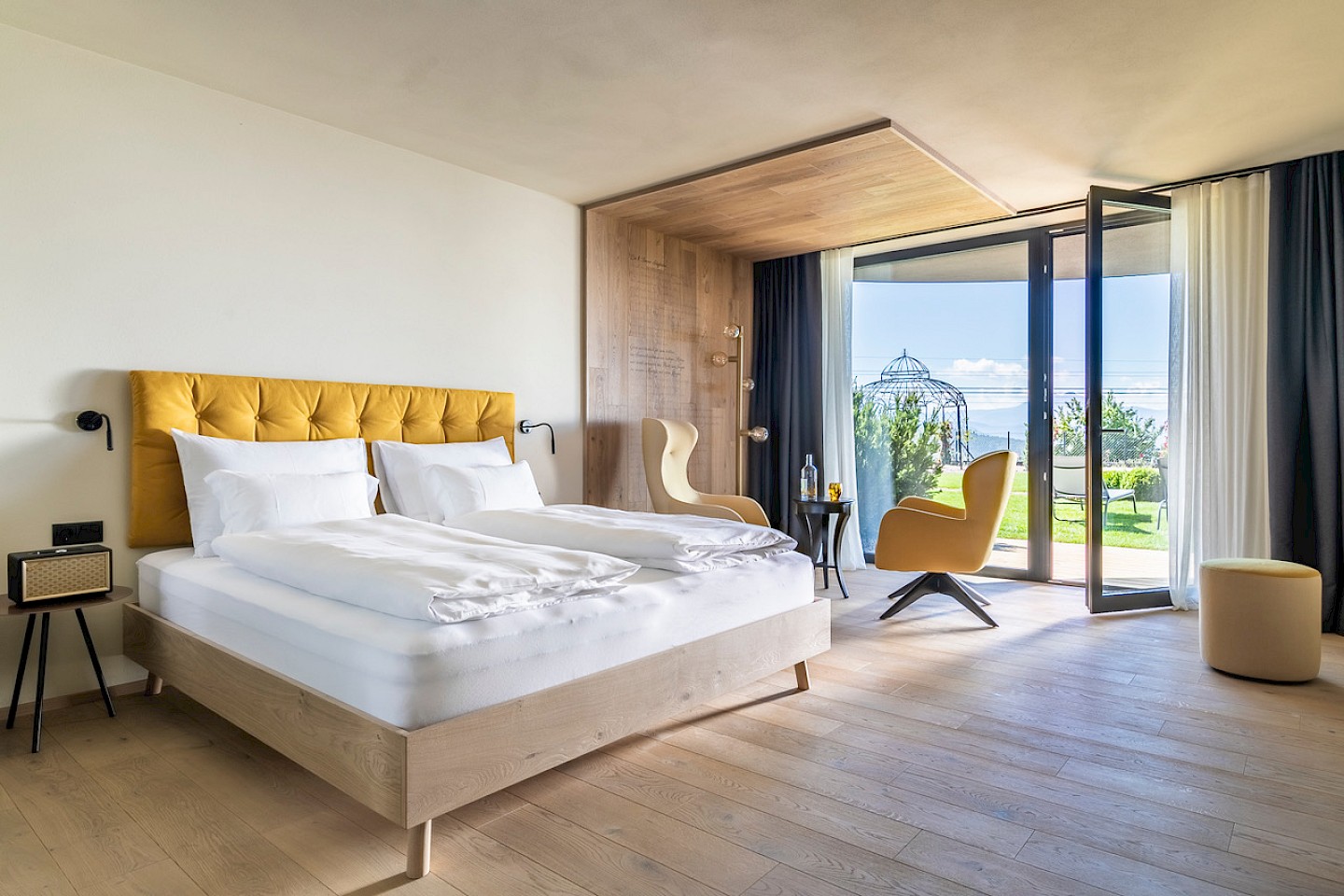 Stadtler Zimmer mit gelbem Kopfteil, Sitzbereich und offener Terrasse mit Blick auf einen Garten.
