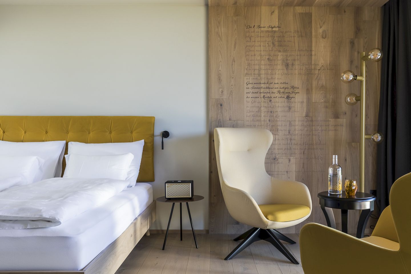 Modernes Hotelzimmer mit gelbem Kopfteil, beigem Sessel, Retro-Radio und hölzerner Akzentwand mit Schrift.