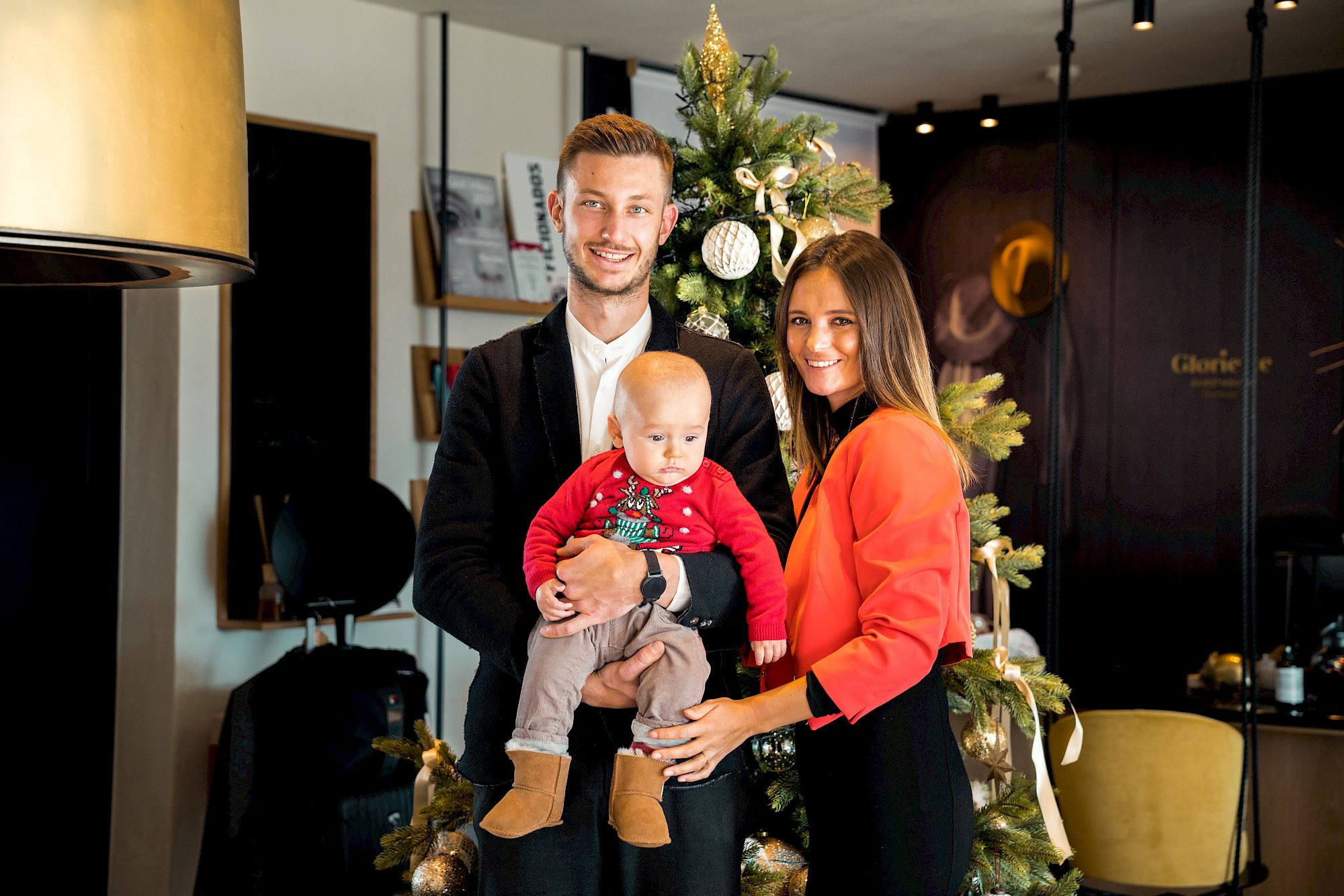 Family smiling in front of a festively decorated Christmas tree. Father holds baby in red sweater.