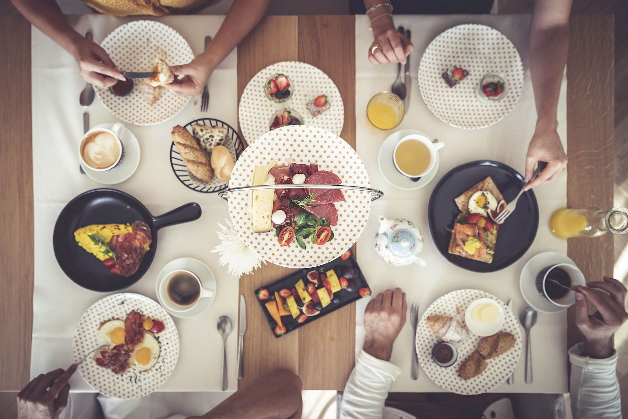 Tavolo colazione con salumi, formaggi, frutta e dolci al Gloriette Guesthouse. Persone intorno al tavolo ben apparecchiato.