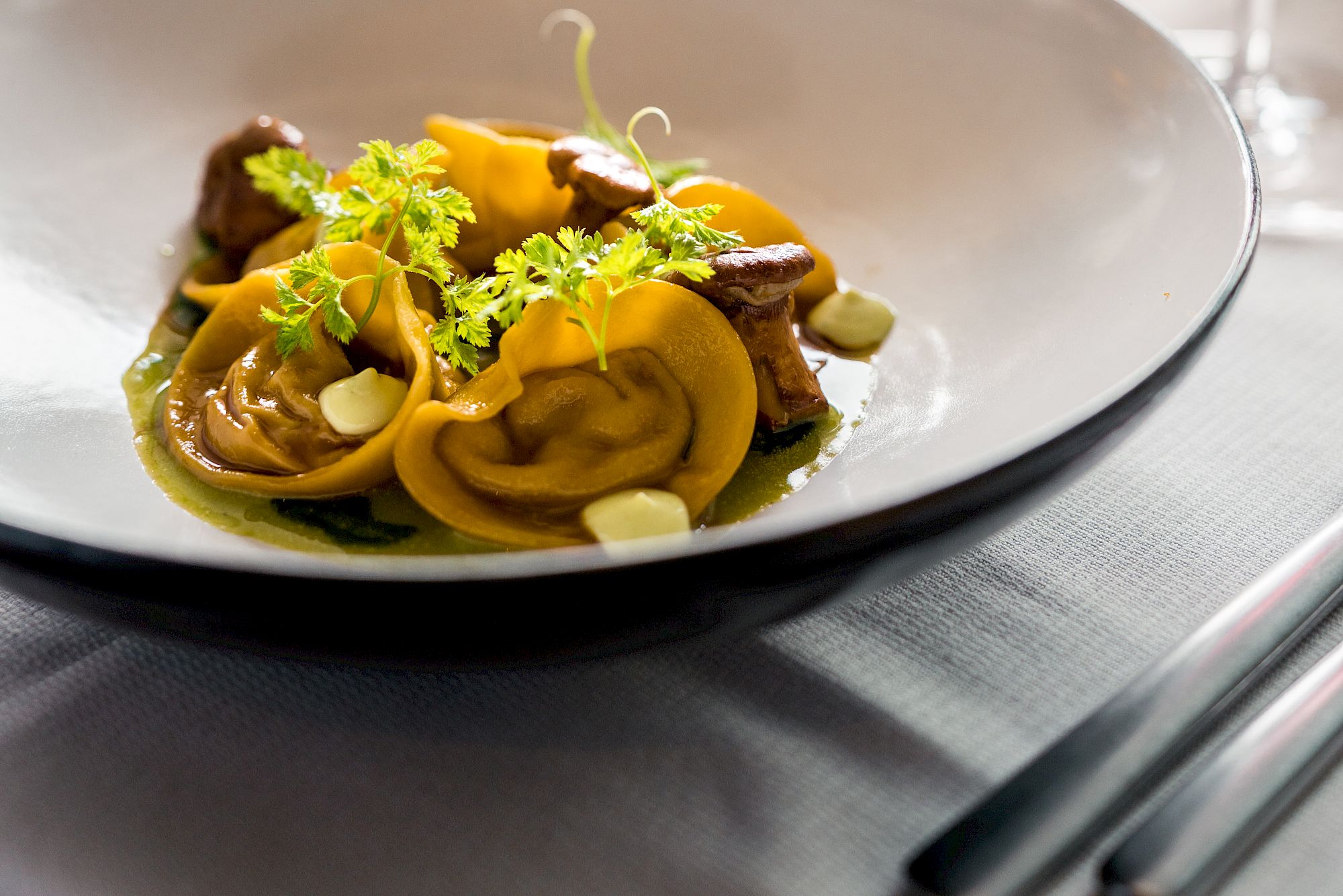 Tortellini mit Soße, dekoriert mit Kräutern, in einem modernen Restaurant serviert, Weingläser im Hintergrund.