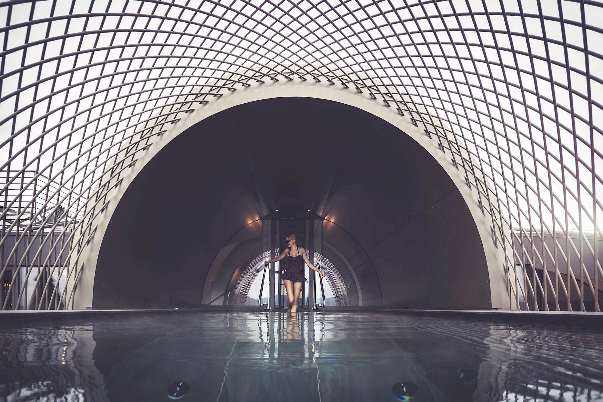 A person stands in the infinity pool below an impressive arched architectural structure.