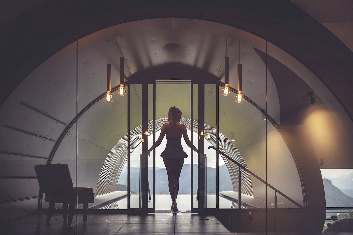 A silhouetted person stands before the infinity pool, overlooking the Renon mountain view. Indoor shot.