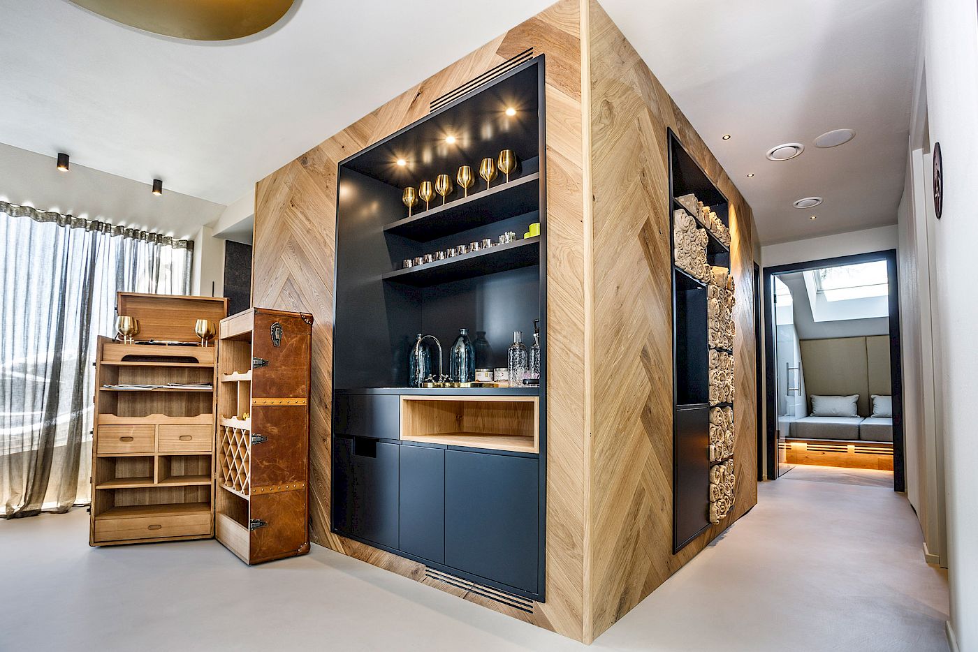 Bar con tè e snack nell´area sauna con parete a scaffali in legno aperto, bicchieri da vino e bottiglie, luce naturale.