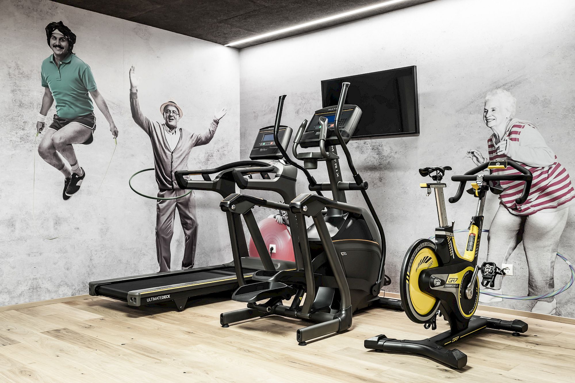 Gym at Gloriette Guesthouse with treadmill, cross trainers, and exercise bike. Wall murals show people jumping and laughing.