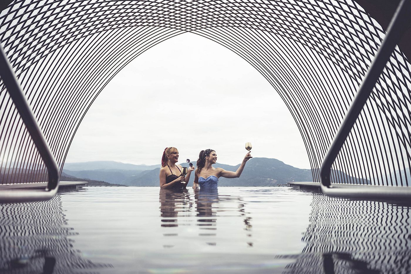 Due donne in un'infinity pool con bicchieri di spumante sotto un arco architettonico.