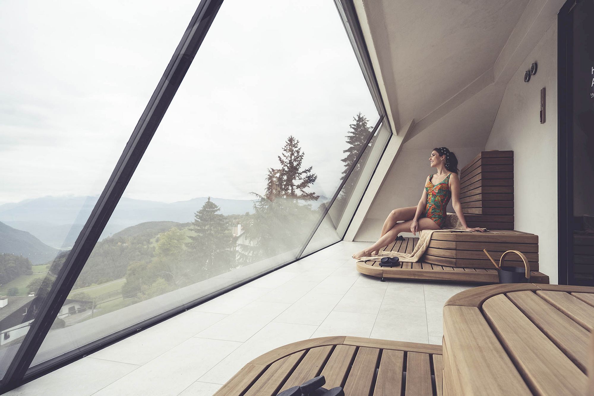 Una donna in sauna moderna con finestra panoramica guarda un paesaggio montano boscoso.