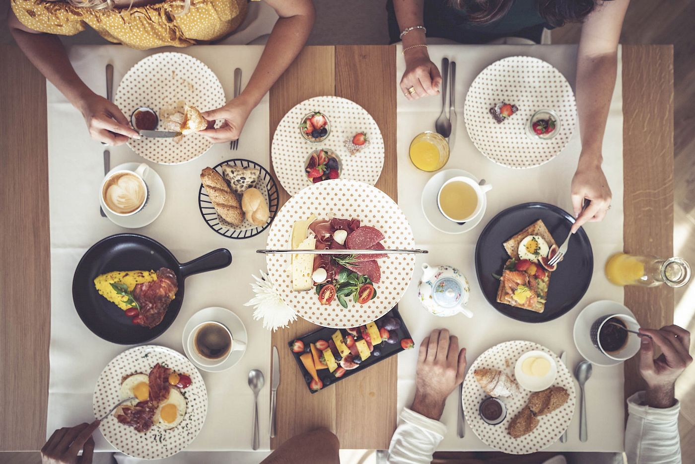 Tavolo colazione con salumi, formaggi, frutta e dolci al Gloriette Guesthouse. Persone intorno al tavolo ben apparecchiato.