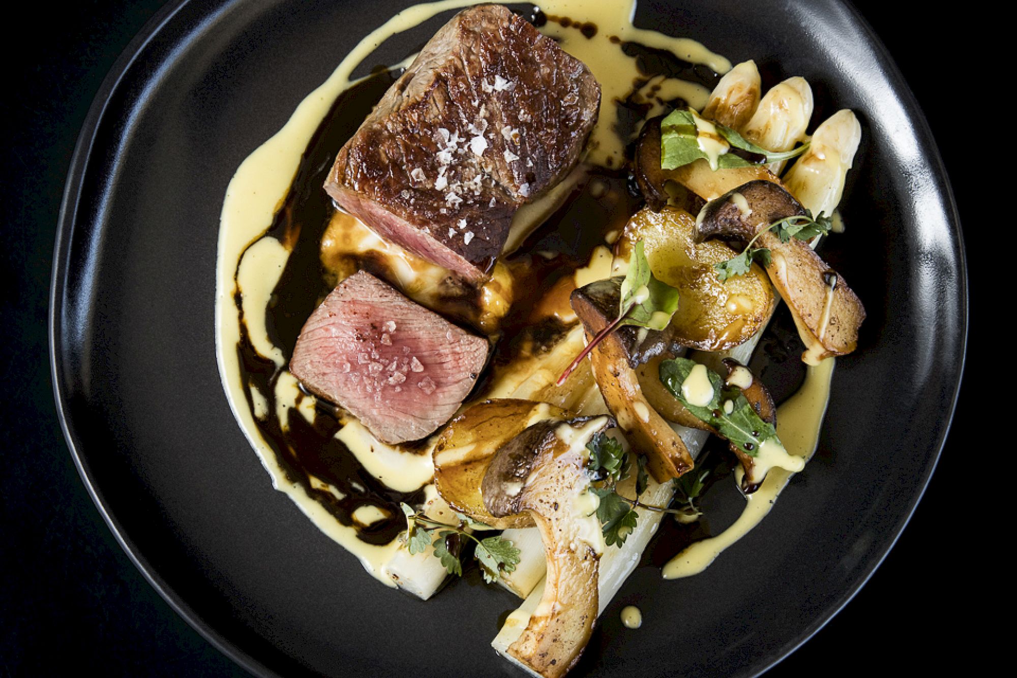 Filet di manzo rosa, asparagi e funghi su piatto nero con salsa cremosa.