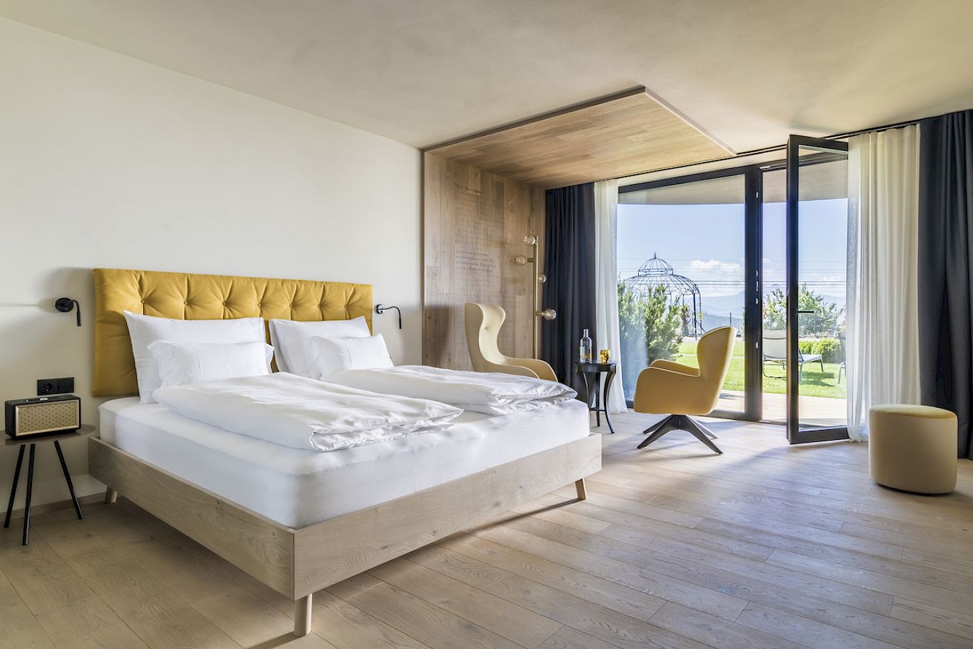 Stadtler Zimmer hotel room with yellow headboard, seating area, and open terrace overlooking a garden.