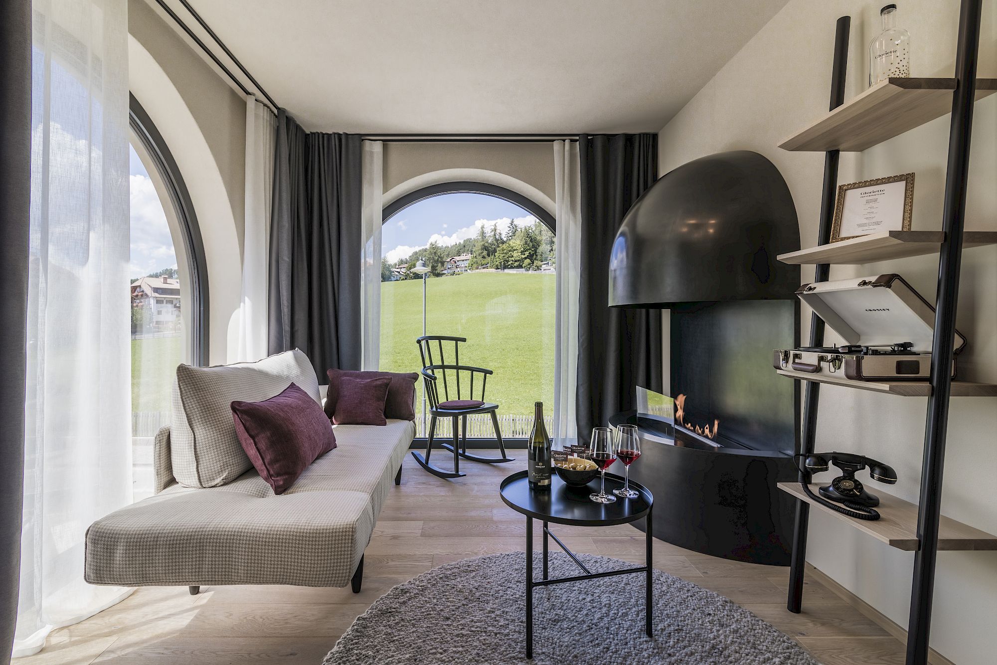Gemütliches Wohnzimmer mit Kamin, Plattenspieler und Ausblick auf eine grüne Wiese durch große Fenster.