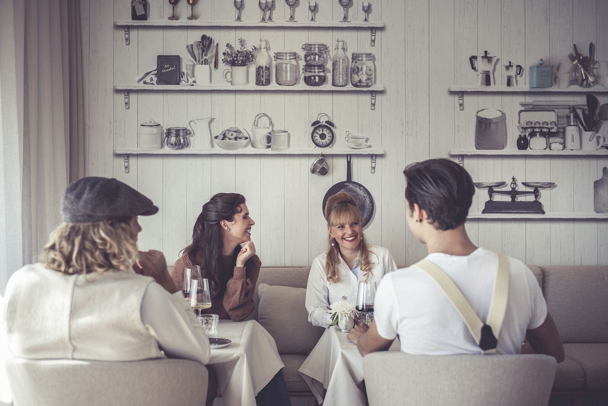 Four people sitting in a cozy café, laughing and drinking wine. Decorative shelves in the background.