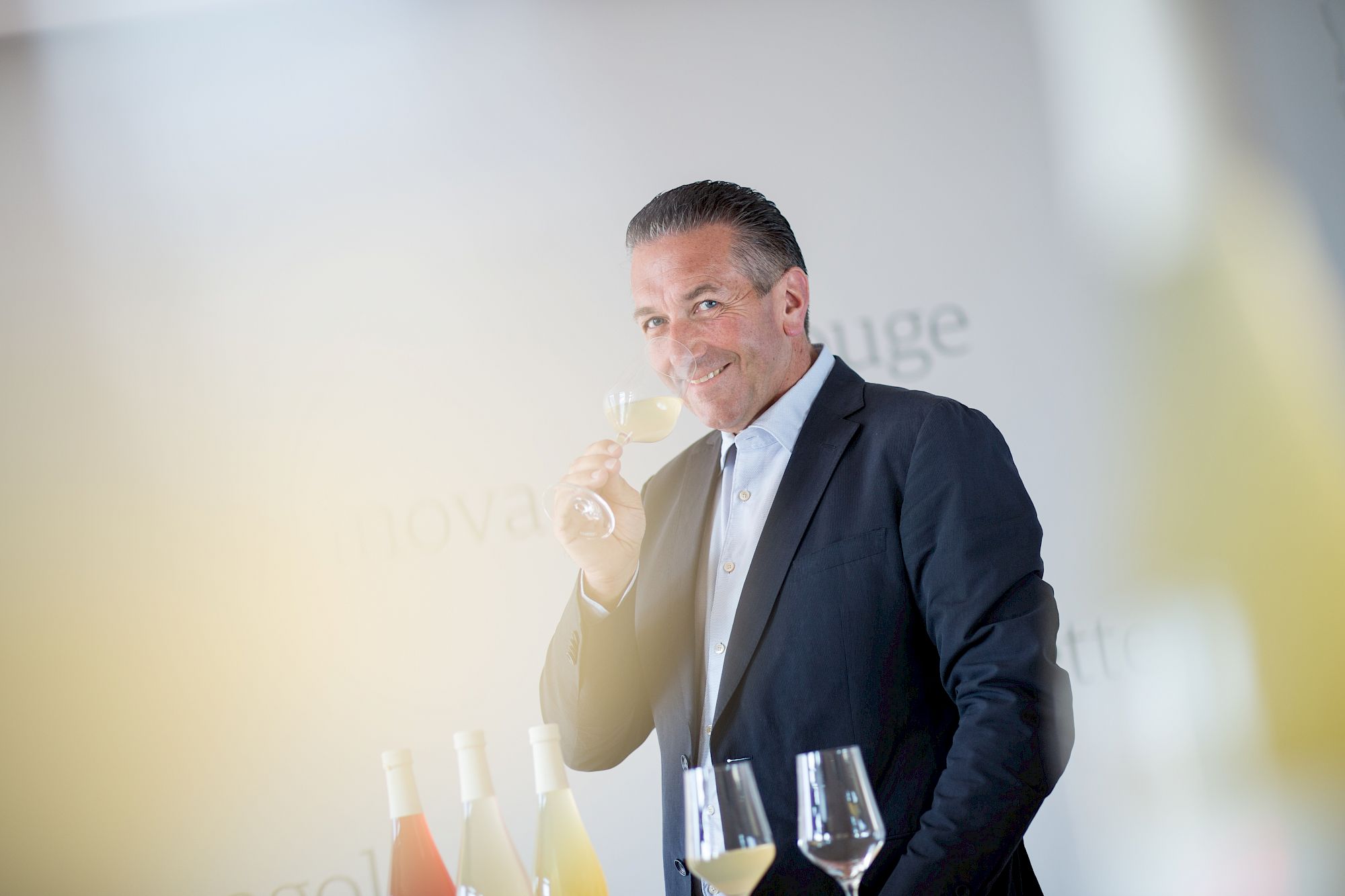 Ein Mann in einem Anzug riecht an einem Glas Weißwein, umgeben von Flaschen auf einem Tisch.
