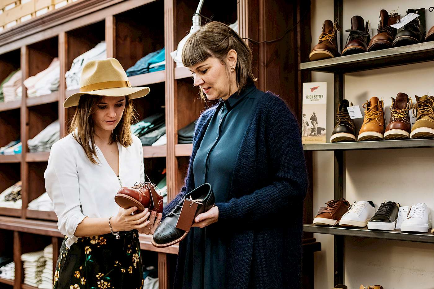 Zwei Frauen betrachten Schuhe in einem Laden; eine trägt einen Hut, die andere hält Schuhe in der Hand.