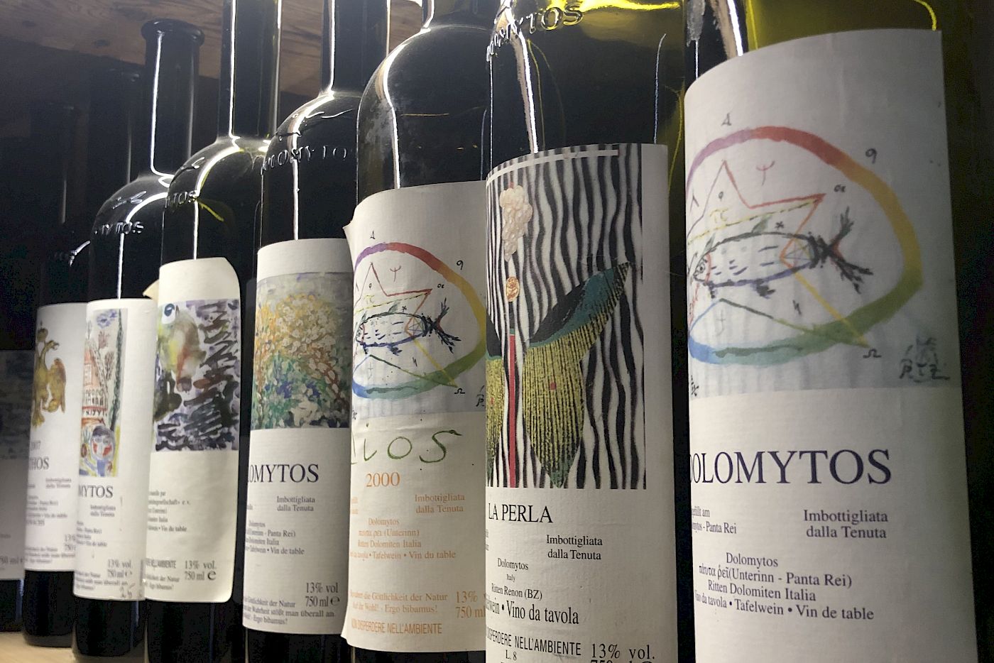 Weinflaschen in einer Regalreihe, verschiedene Etiketten mit bunten Designs. Beleuchtung von oben.