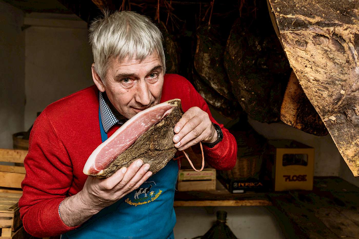Ein Mann in einer Fleischerei riecht an einem großen Stück Schinken. Holzregale sind im Hintergrund sichtbar.