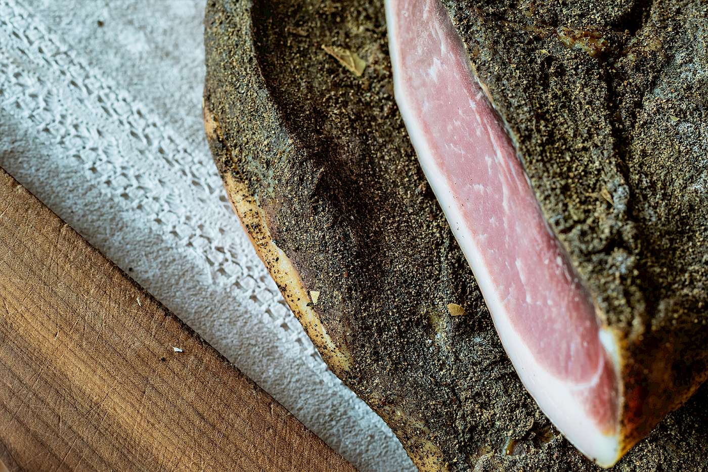 Ein Stück Rohschinken mit Kräutern und Gewürzen auf einem Holzbrett und einer Spitzen-Tischdecke.