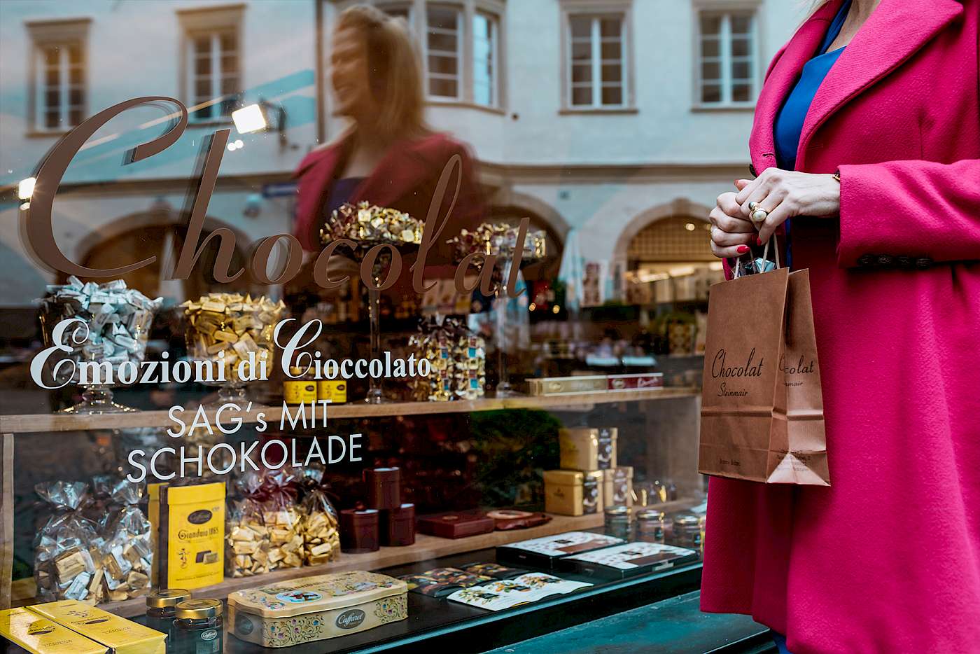 Frau in rotem Mantel hält Papiertüte vor Schaufenster eines Schokoladengeschäfts mit verzierten Schachteln.