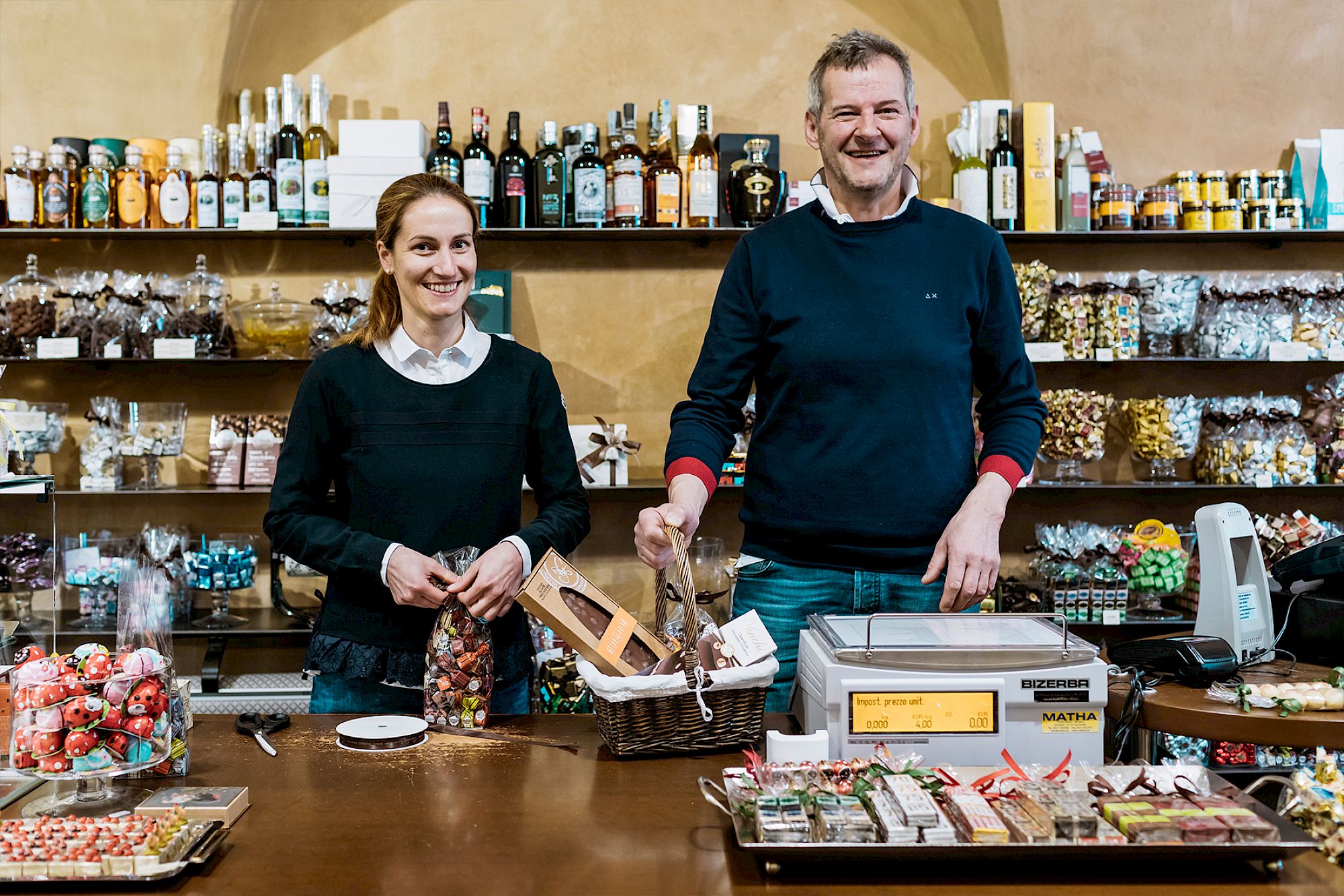 Zwei Personen stehen in einem Feinkostladen und lächeln hinter einem Verkaufstresen voller Produkte.