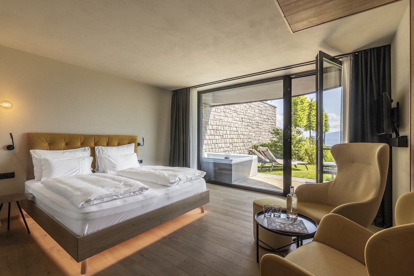 Garten Whirlpool Suite mit Doppelbett, bequemen Sesseln und großem Fenster mit Blick auf Terrasse und Natur.