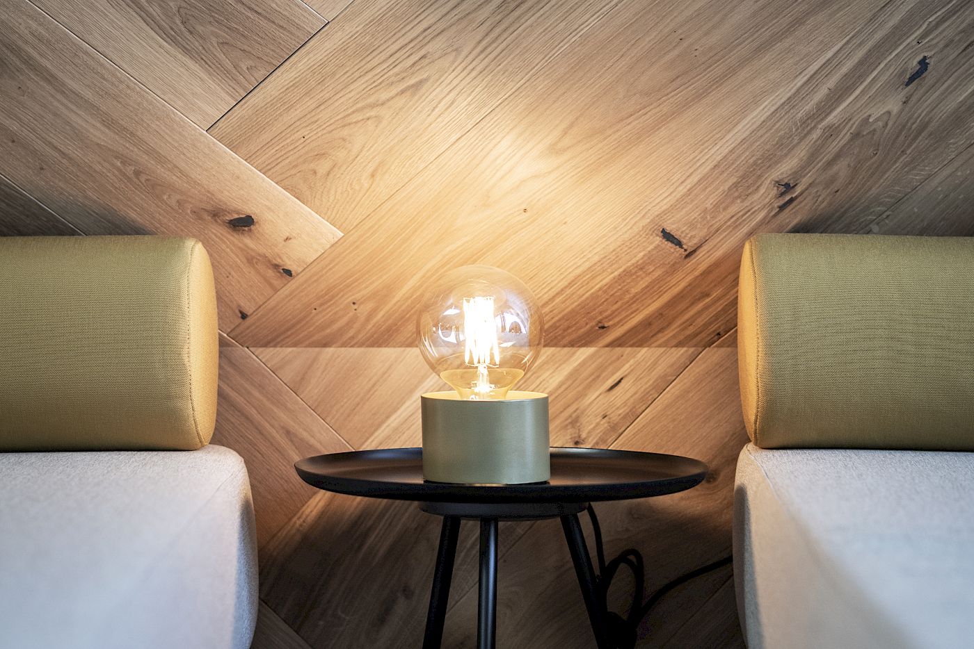 Two beds with a bedside lamp on a small table, in front of a wooden paneled wall.
