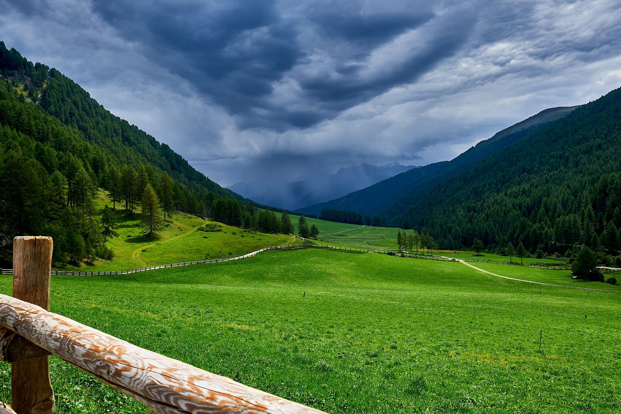 Prato alpino verde in una valle con una staccionata in legno in primo piano e cielo nuvoloso.