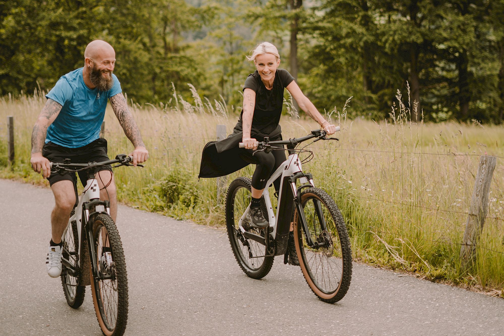 Due persone pedalano su una strada di campagna, circondate da alberi. Entrambi sorridono.