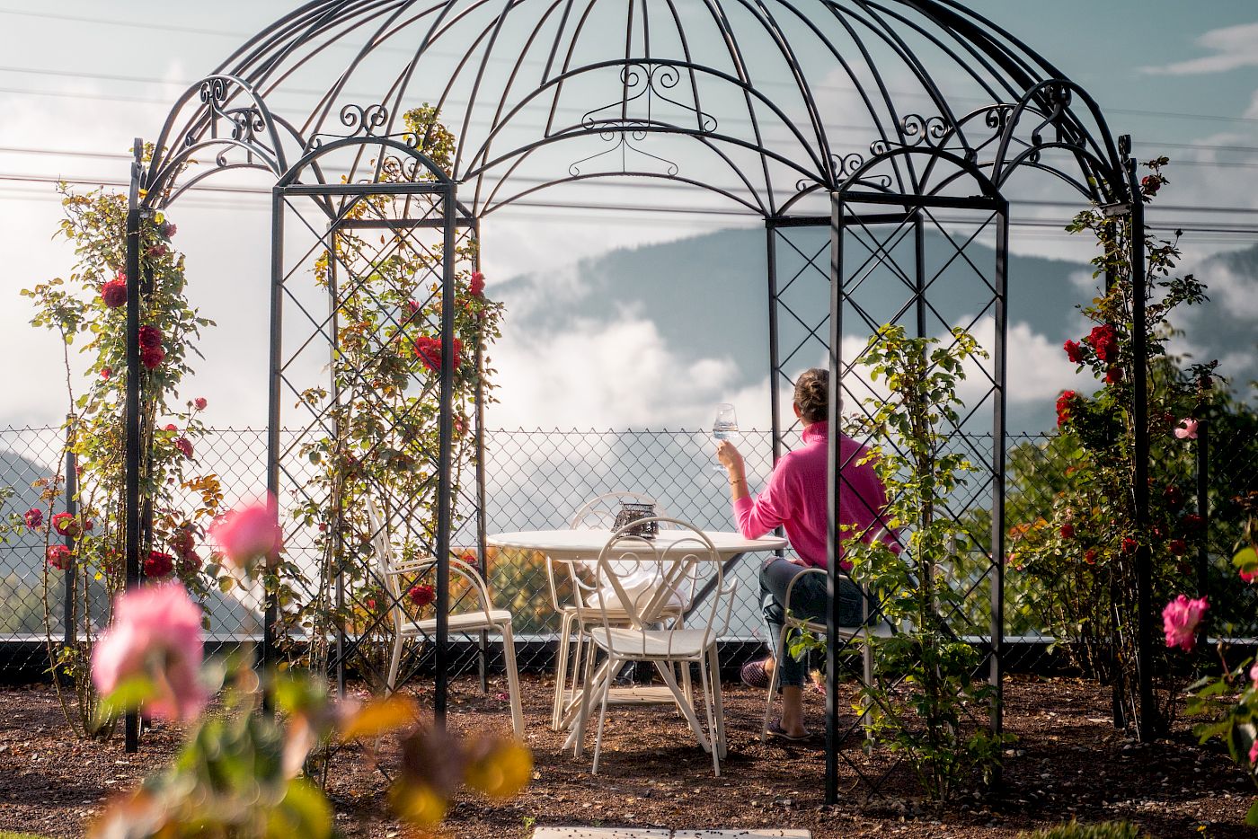 Frau in rosa Jacke sitzt unter Pavillon im Garten vom Gloriette Guesthouse, trinkt Kaffee mit Rosenausblick.