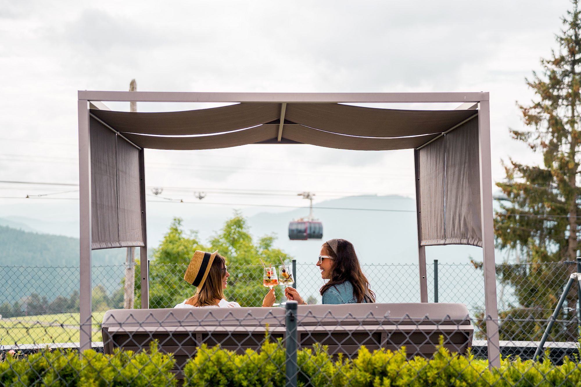 Two people sit in a pavilion, clinking drinks, as a cable car passes by in the background.