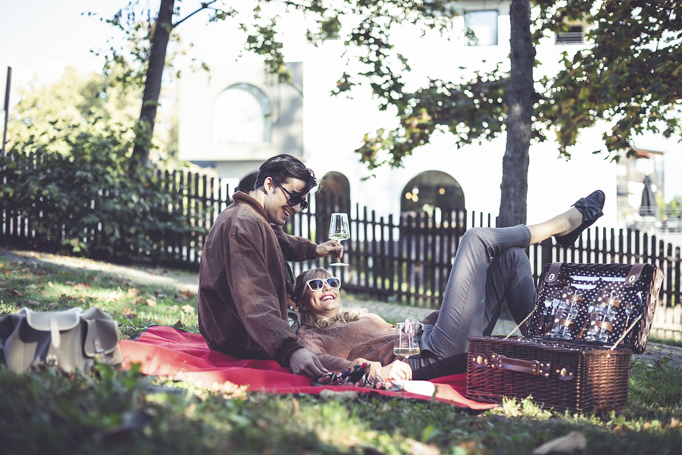 Ein Paar genießt ein Picknick im Park, sitzt auf einer Decke, trinkt Wein, umgeben von Bäumen.