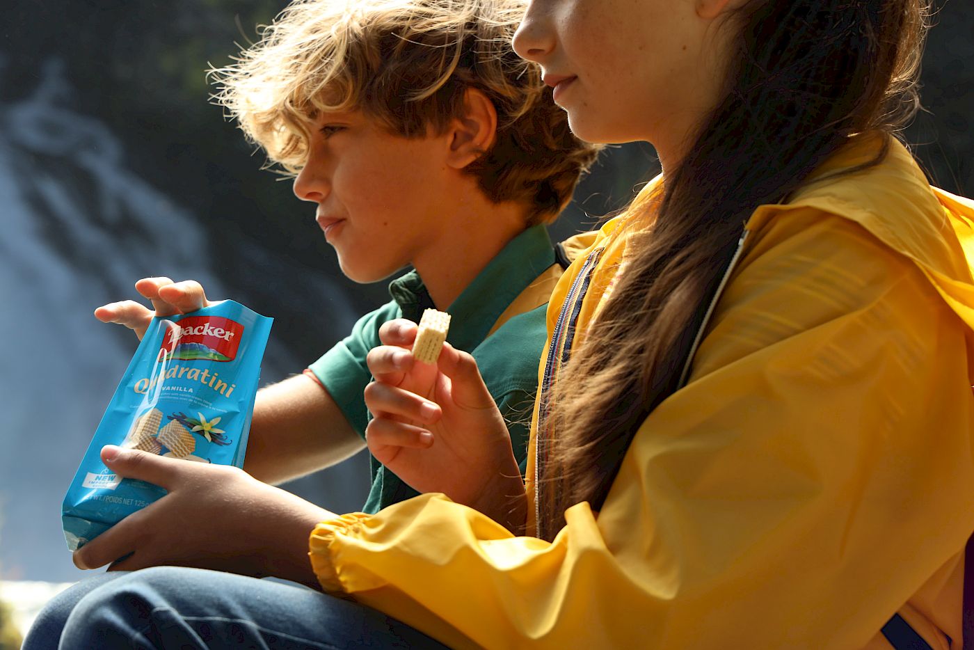 Zwei Kinder sitzen vor einem Wasserfall und essen gemeinsam Waffeln aus einer blauen Packung.