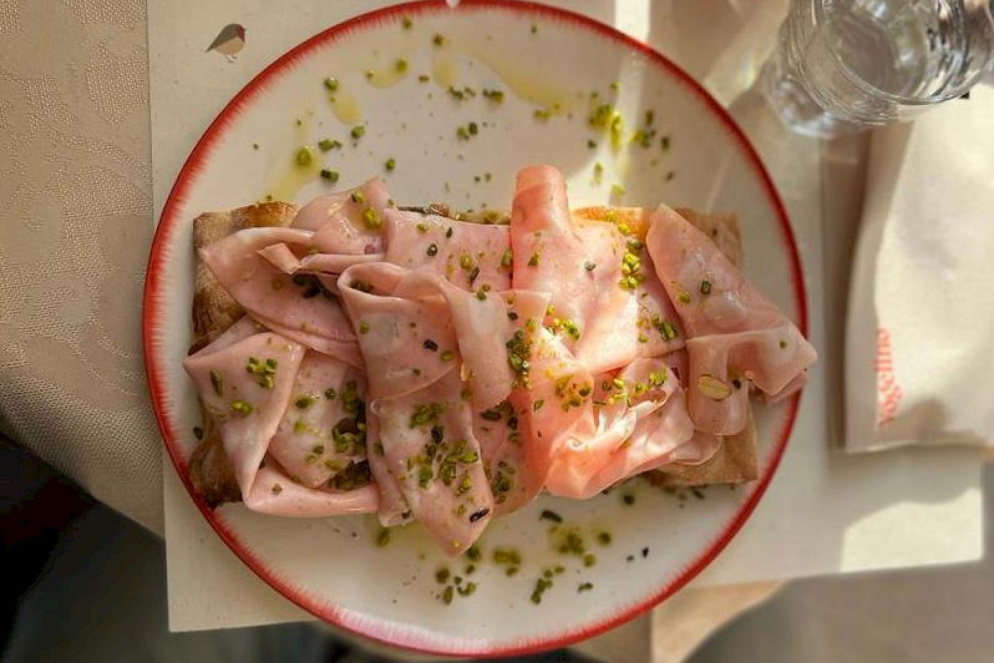 A plate with a slice of bread, topped with mortadella and herbs, on a table.