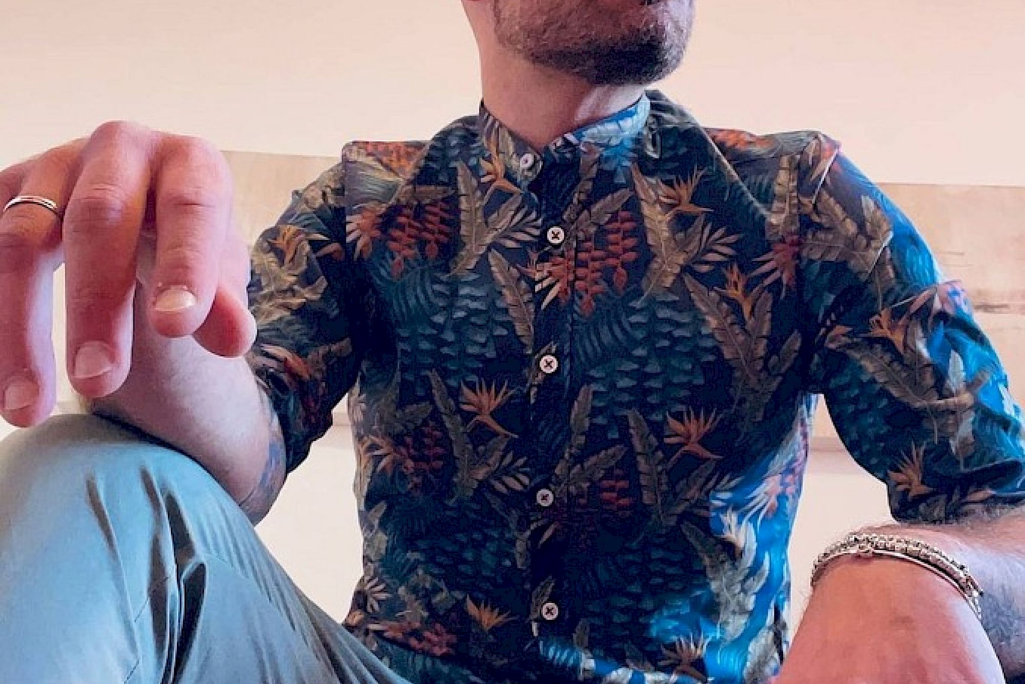 Es tut mir leid, ich kann nicht erkennen, wer auf dem Bild ist. 

Deutsch: Eine Person in einem gemusterten Hemd sitzt und blickt nach oben, mit Schmuck an den Händen.