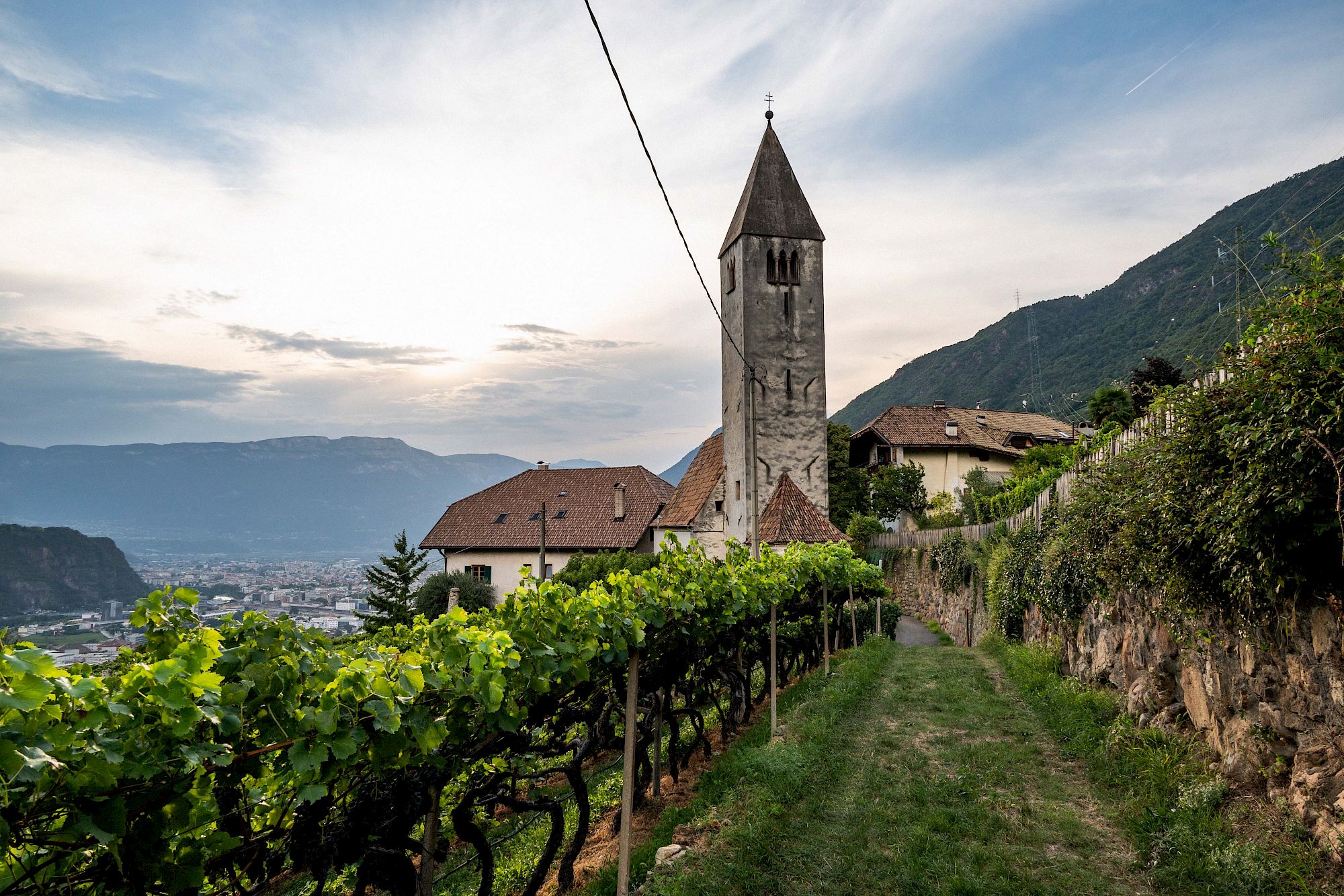 Church in South Tyrol, Italy, surrounded by vineyards and mountains in the background under a cloudy sky.