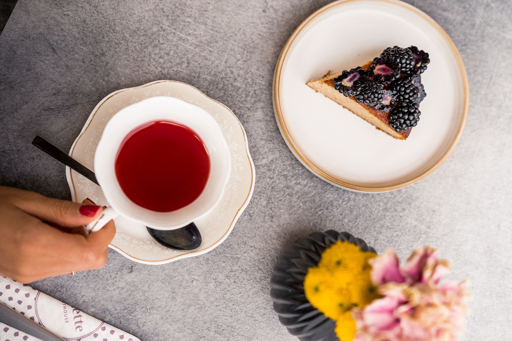 Tazza di tè rosso e pezzo di torta alle more su piatto. Tavolo con superficie grigia e vaso di fiori rosa e gialli.