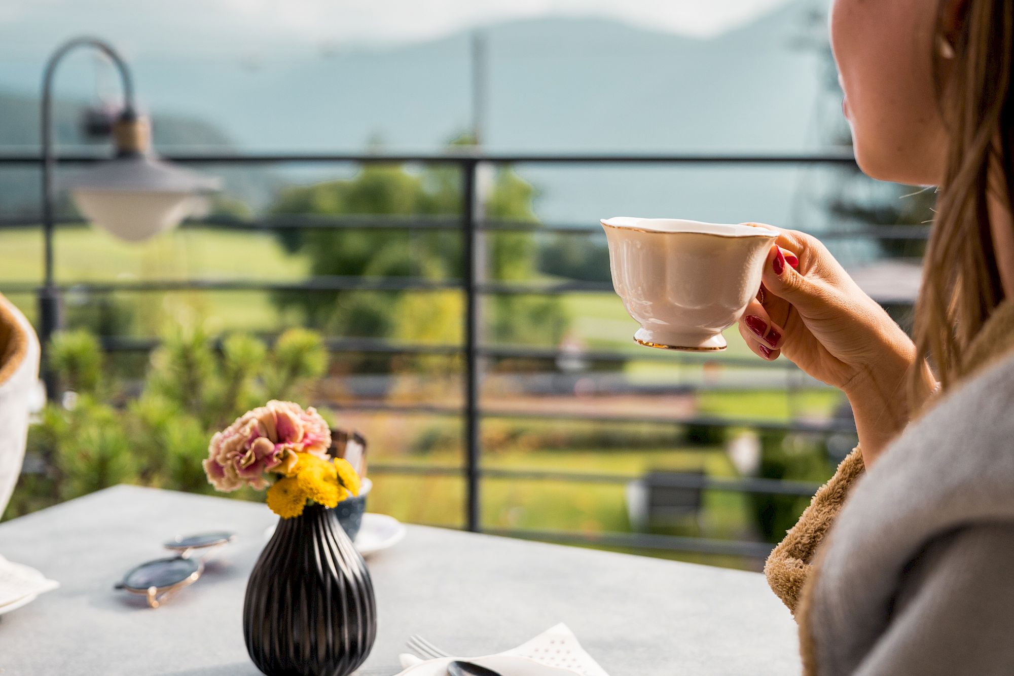 Donna gusta un caffè sulla terrazza del Gloriette Guesthouse con vista sui monti, accanto un piatto di torta e un vaso di fiori.