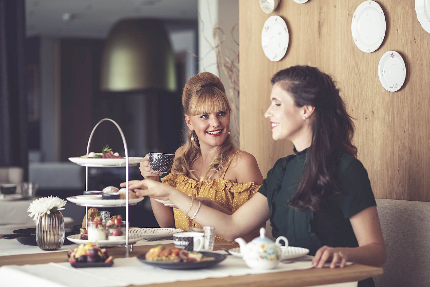 Two women enjoy tea and pastries at Gloriette Guesthouse with decorative plates on the wall.